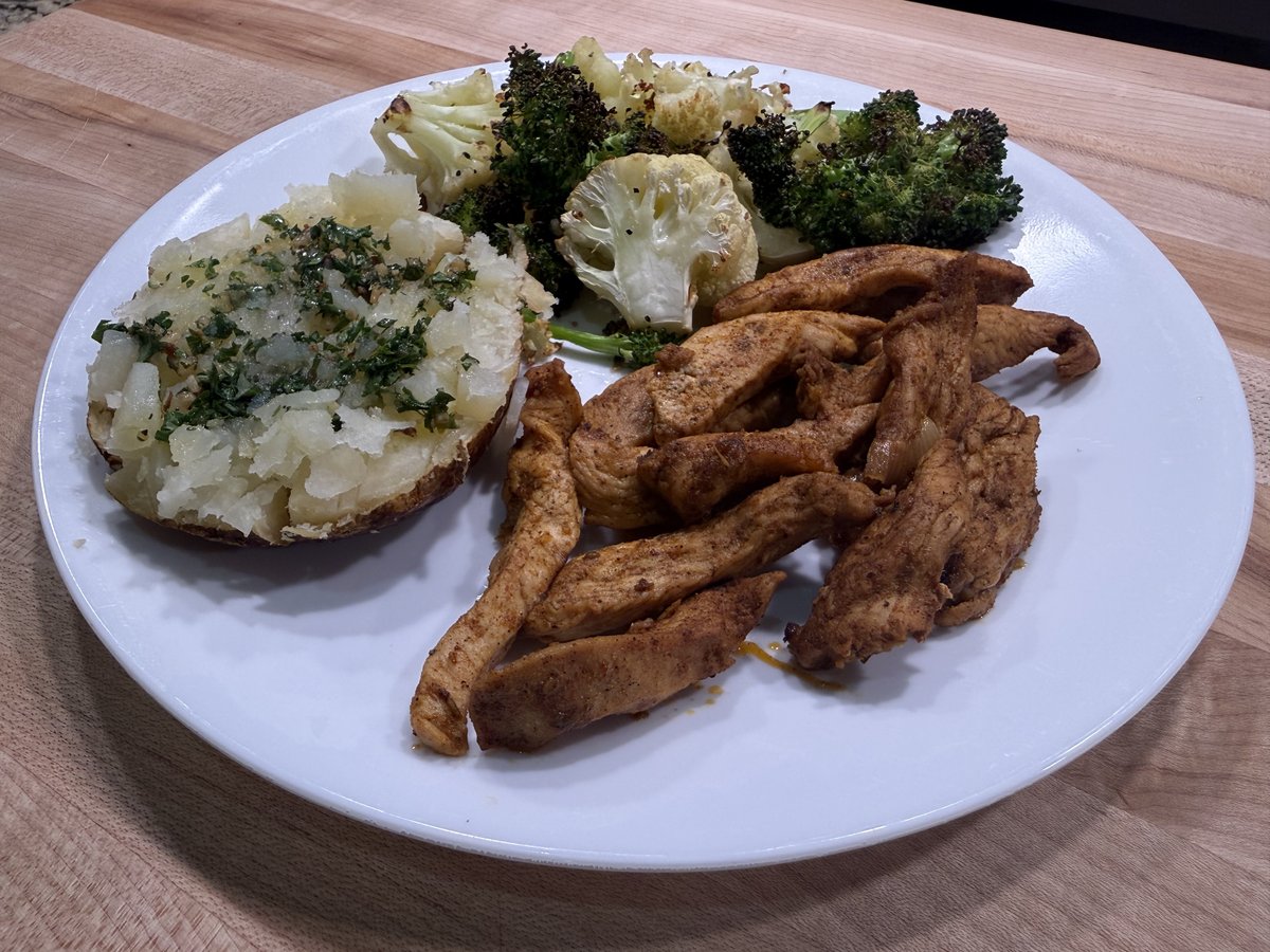 Dinner plate: grilled chicken breast strips, half a baked potato topped with herbs, and roasted broccoli and cauliflower